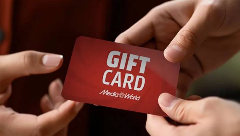 Una mano tiene una carta regalo rossa con scritte bianche, mentre un'altra mano tiene un telefono.