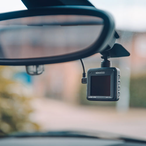 Specchietto retrovisore auto con dash cam Kenwood nera montata. Cavo nero visibile. Sfondo sfocato di alberi esterni.