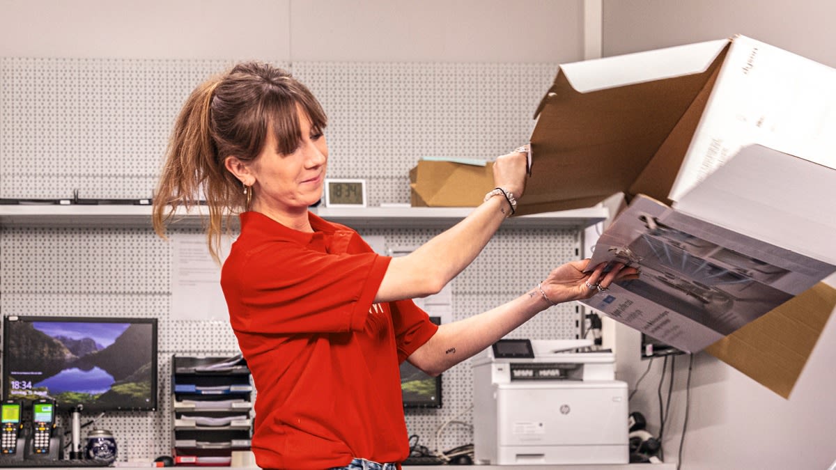 Donna in maglietta rossa apre scatola con foto. Ufficio con schermo, stampante e scaffali.