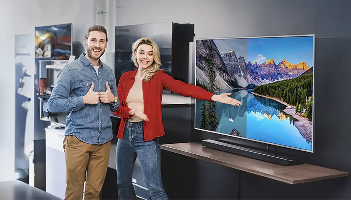 Uomo e donna davanti a TV che mostra montagne e lago su una mensola. L'uomo fa il gesto del pollice in su.