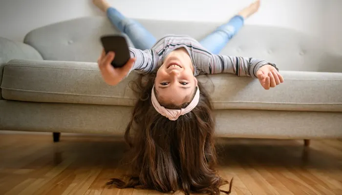 Una persona sorridente è sdraiata a testa in giù su un divano grigio mentre tiene in mano un telecomando, con i capelli lunghi che toccano il pavimento in legno.
