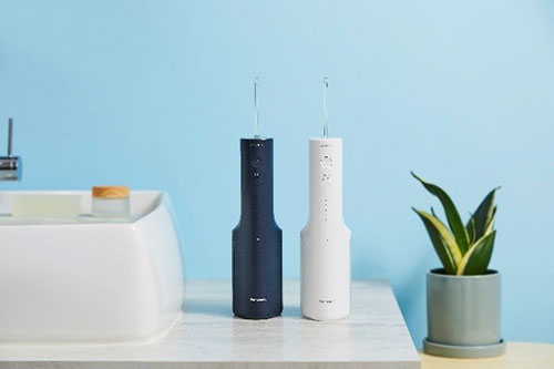 Due irrigatori orali, uno blu e uno bianco, su un ripiano accanto a un lavandino bianco e una pianta verde in vaso grigio.