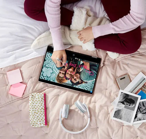 Donna seduta sul letto usa un tablet con foto di gruppo. Ci sono cuffie, foto, note adesive e una pochette sul letto.