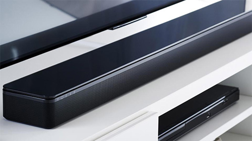 Barra audio nera su ripiano bianco, con un lettore DVD nero sottostante. TV visibile in alto.