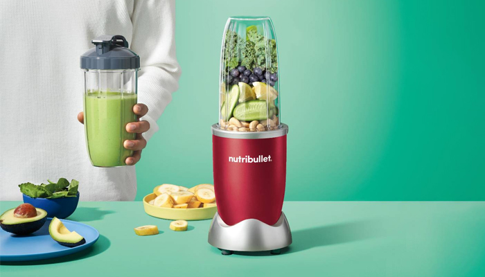 Un frullatore Nutribullet rosso con frutta e verdura. Una persona tiene in mano un bicchiere di frullato verde. Avocado e banane sul tavolo.