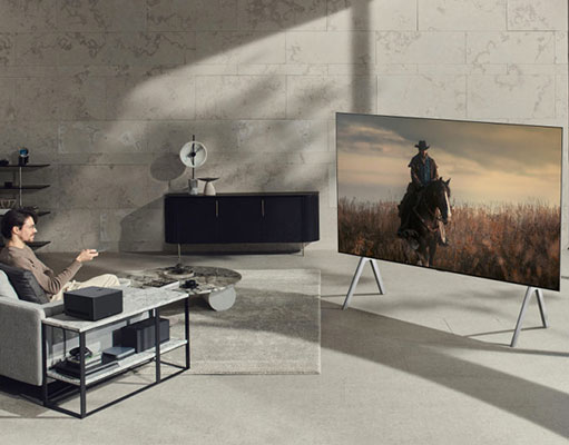 Soggiorno con uomo su divano grigio, tavolino con oggetti neri e ripiani. TV mostra cavaliere in campo. Mobiletto nero e ventola sullo sfondo.