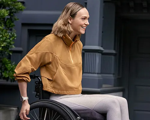 Donna sorridente su sedia a rotelle, indossa felpa ocra e pantaloni chiari. Dietro, edificio grigio scuro e vegetazione.