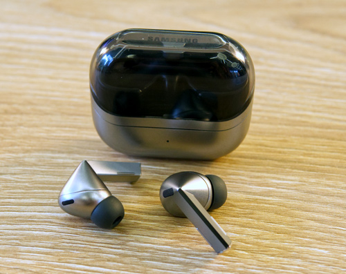 Custodia nera e argento per auricolari wireless, con due auricolari argento e nero su superficie di legno chiara.