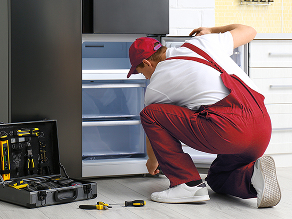 Un riparatore in tuta rossa esamina un frigorifero aperto con una cassetta degli attrezzi e cacciaviti sul pavimento.