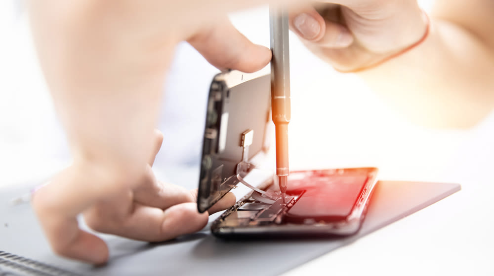 Due mani riparano un telefono aperto con un cacciavite su una superficie grigia.