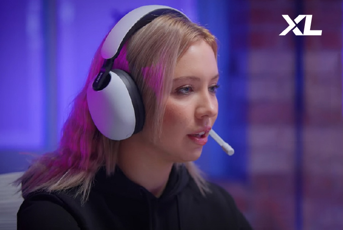 Donna con cuffie bianche e microfono indossa una felpa nera, sfondo blu e scritta "XL".