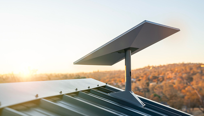 Pannello Starlink grigio su tetto metallico, con supporto. Sfondo sfocato di alberi e cielo chiaro al tramonto.