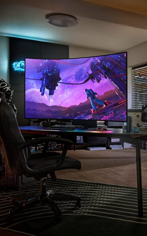 Ampio schermo curvo su scrivania con sedia da gioco. Lo schermo mostra un paesaggio viola fantastico. Tappeto a righe sul pavimento.