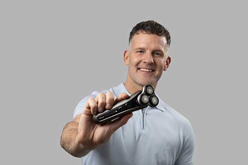 Uomo sorridente che tiene un rasoio elettrico nero e argento su sfondo grigio. Indossa una polo chiara.