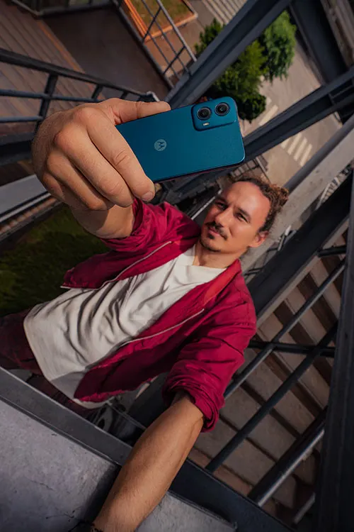 Un uomo con giacca rossa fa un selfie con un telefono blu; sullo sfondo, un edificio e vegetazione.