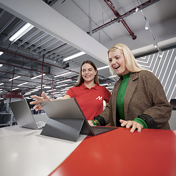 Due donne guardano un portatile aperto su un tavolo rosso. La donna a sinistra indossa una maglietta rossa, l'altra una giacca marrone.