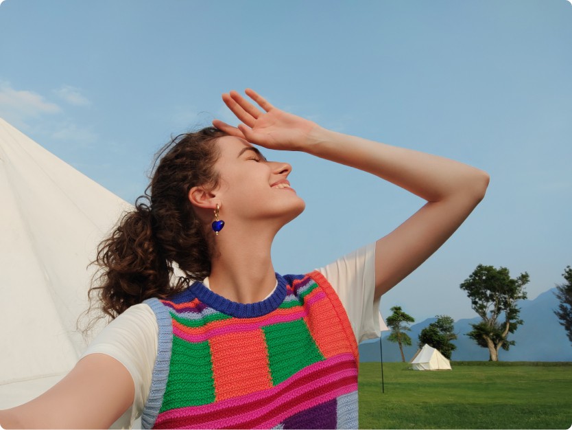 Donna sorride, mano sulla fronte, in un campo. Tenda bianca a sinistra e una sullo sfondo, alberi e cielo azzurro. Indossa orecchini blu.