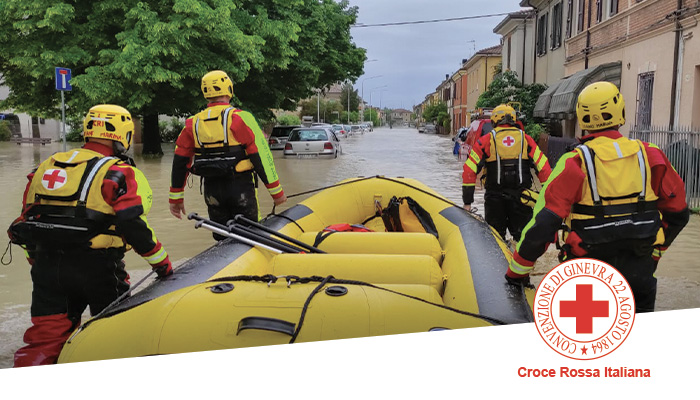 Soccorritori in giallo e rosso spingono gommone giallo in strada allagata. Auto e case sullo sfondo.