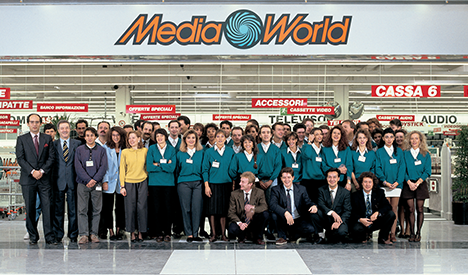 Un folto gruppo di persone posa davanti al negozio MediaWorld. Insegne rosse indicano "Accessori", "Televisori", "Audio" e "Cassa 6".