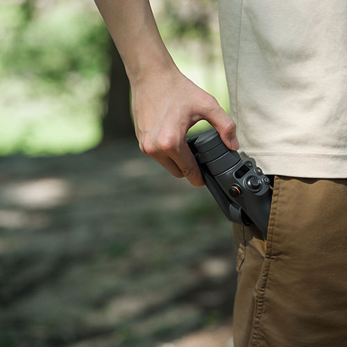 Uomo con maglietta beige e pantaloni marroni. Tiene in mano un gimbal nero che sporge dalla tasca. Sfondo verde sfocato.