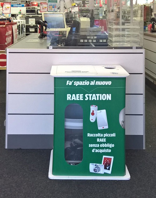 Cassa bianca con postazione verde "RAEE Station" per raccolta rifiuti elettronici. Sullo sfondo, scaffali di un negozio.