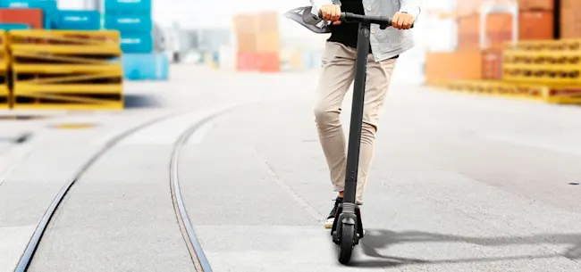 Persona su monopattino elettrico su strada con rotaie, contenitori sullo sfondo.