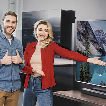 Uomo in jeans blu e donna in giacca rossa accanto a TV con paesaggio montano. Entrambi sorridono e indicano lo schermo.