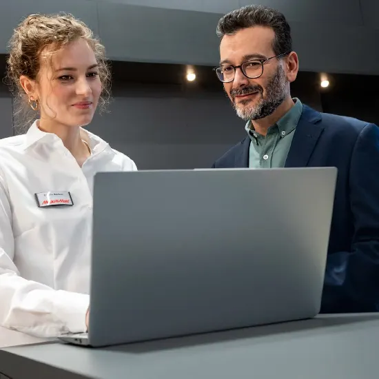 Una donna e un uomo guardano un laptop grigio su un tavolo grigio. La donna indossa una camicia bianca e un cartellino identificativo.