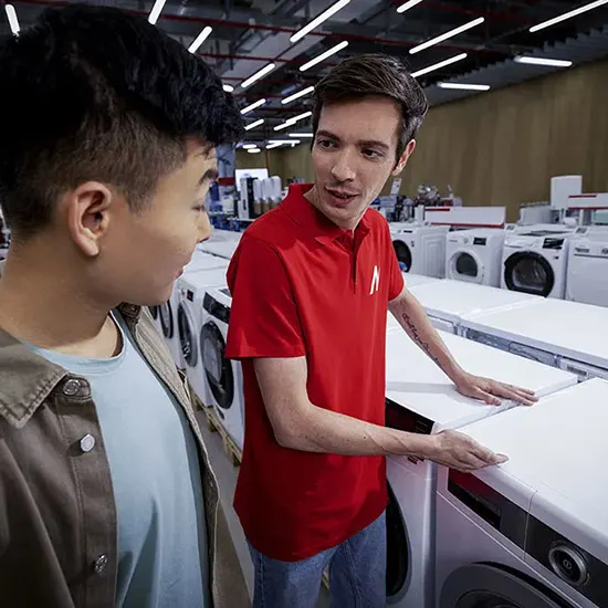 Un venditore in polo rossa mostra lavatrici bianche a un cliente in camicia azzurra, in un negozio illuminato.