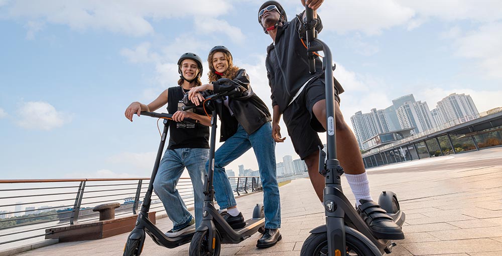 Tre persone con caschi su monopattini neri, con edifici sullo sfondo, su un ponte.