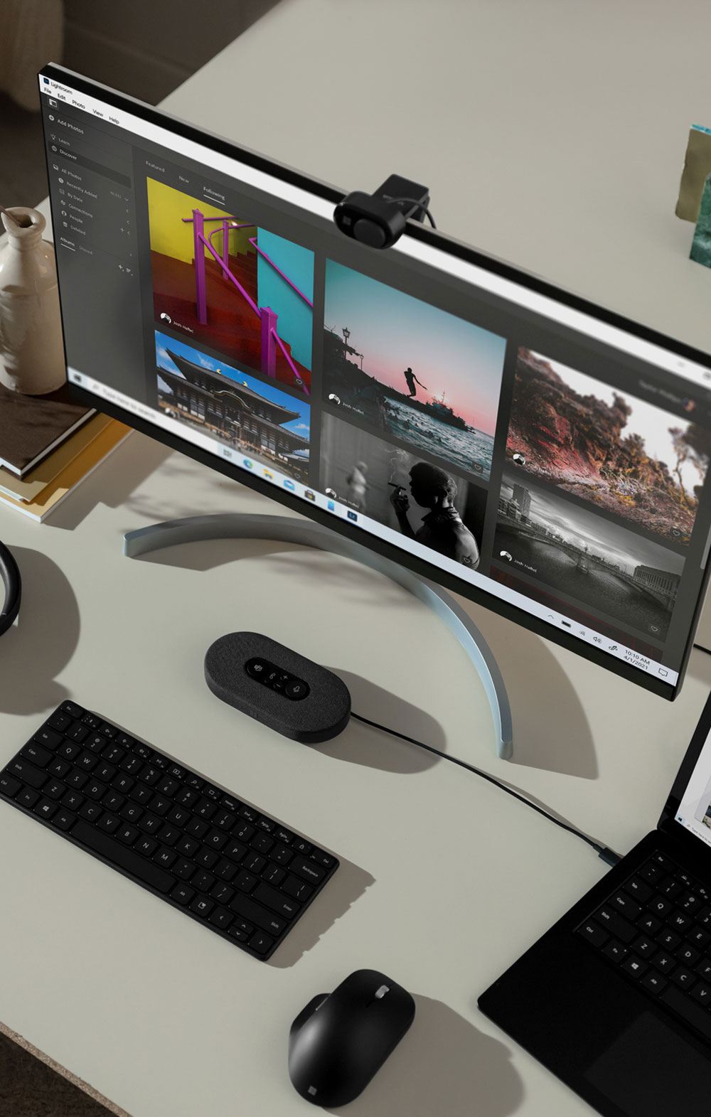 Scrivania con monitor, webcam, tastiera nera, mouse, laptop e dispositivo di controllo. Sullo schermo appaiono diverse foto.