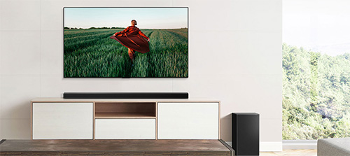 Mobiletto TV bianco con soundbar. Sopra, schermo piatto che mostra una persona con carriola in un campo verde.