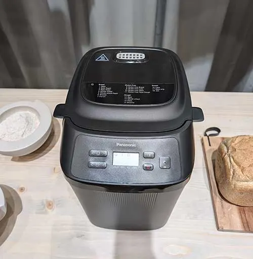 Macchina del pane nera su tavolo di legno chiaro, accanto a ciotola di farina e tagliere con pane. Tende chiare sullo sfondo.