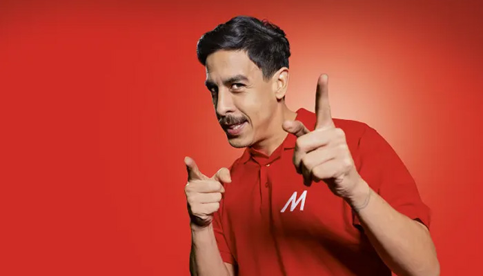 Uomo in polo rossa con lettera "M", indica davanti a sfondo rosso, sorridendo.