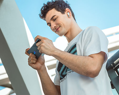 Uomo con auricolare bianco, maglietta bianca, tiene in orizzontale un telefono grigio. Cielo azzurro sullo sfondo.