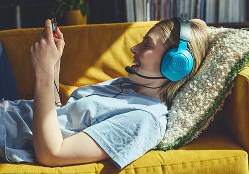Donna distesa su divano giallo con cuffie azzurre, microfono e cuscino beige, che usa dispositivo elettronico. Sullo sfondo, libreria.