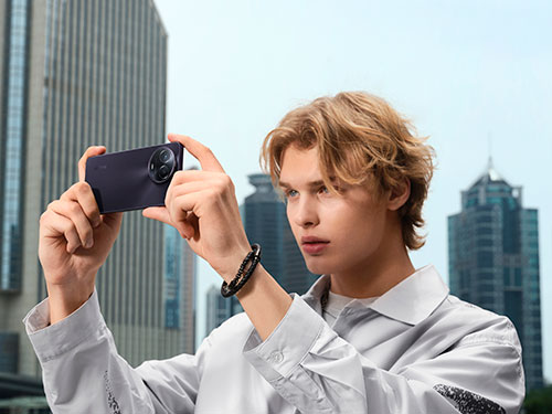 Uomo tiene un telefono nero orizzontalmente, sullo sfondo palazzi grigi e cielo chiaro. Indossa una camicia bianca e un braccialetto scuro.