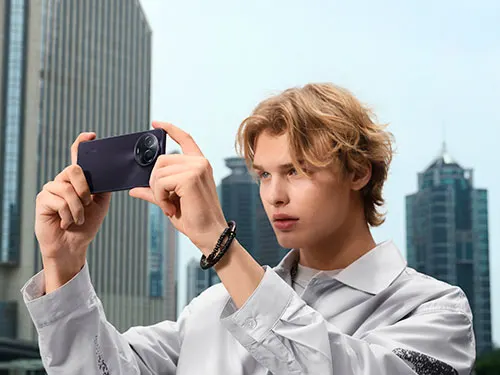 Uomo tiene un telefono nero orizzontalmente, sullo sfondo palazzi grigi e cielo chiaro. Indossa una camicia bianca e un braccialetto scuro.