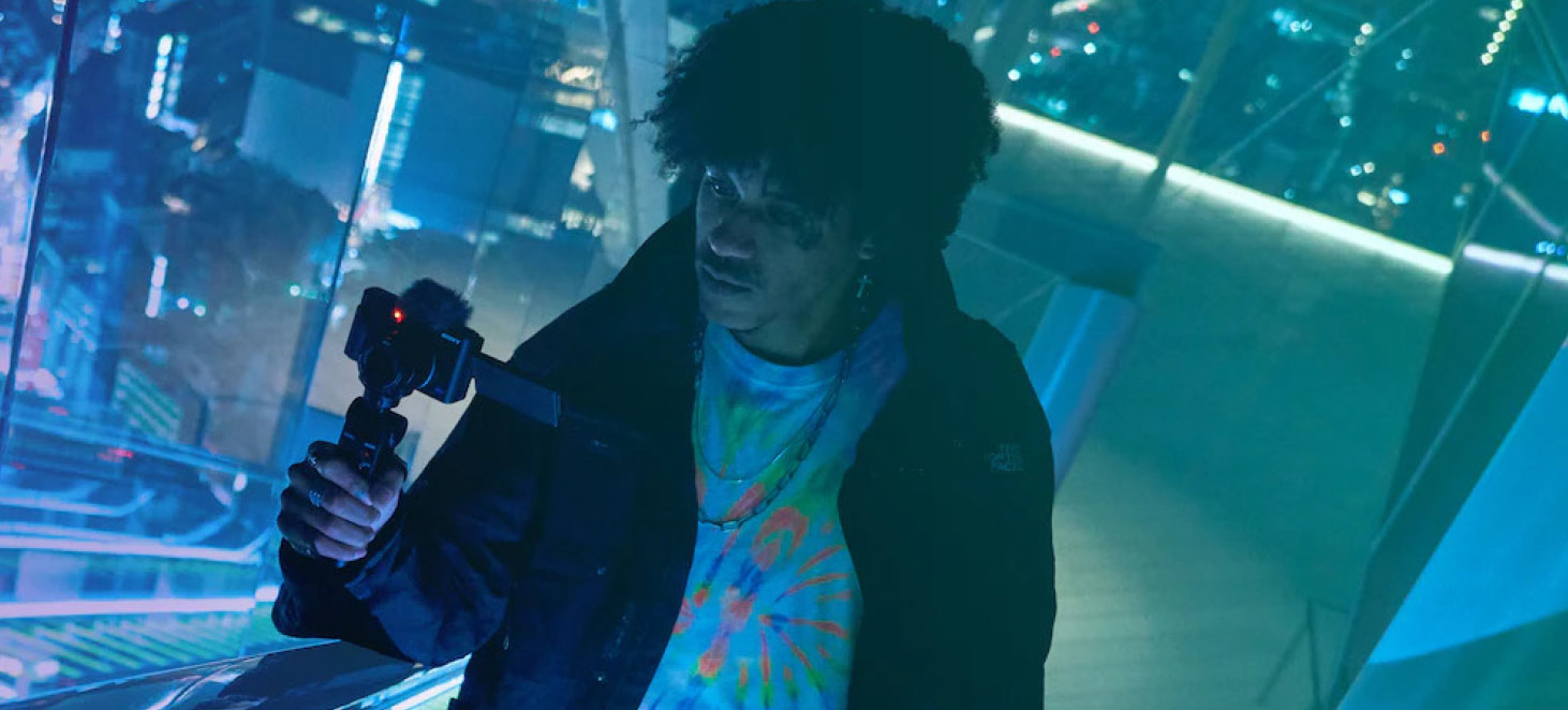 Un uomo riprende con una videocamera in un ambiente urbano futuristico illuminato di blu. Indossa una maglietta colorata e una giacca scura.