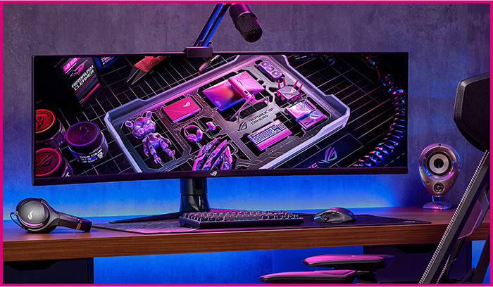 Cuffie, tastiera e mouse su scrivania di legno. Monitor con immagine di accessori tecnologici. Microfono e altoparlante visibili.