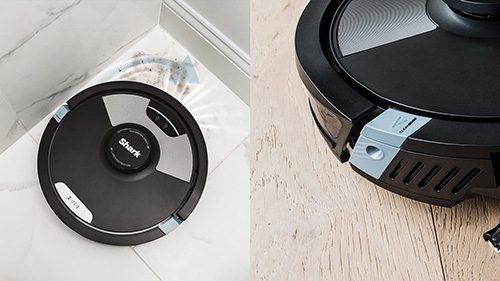 Un aspirapolvere robot nero e grigio si trova su un pavimento bianco e un pavimento in legno chiaro.