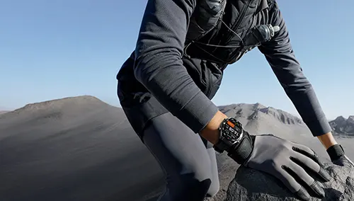 Escursionista con guanti tocca roccia. Indossa orologio e gilet. Montagne grigie e cielo azzurro sullo sfondo.