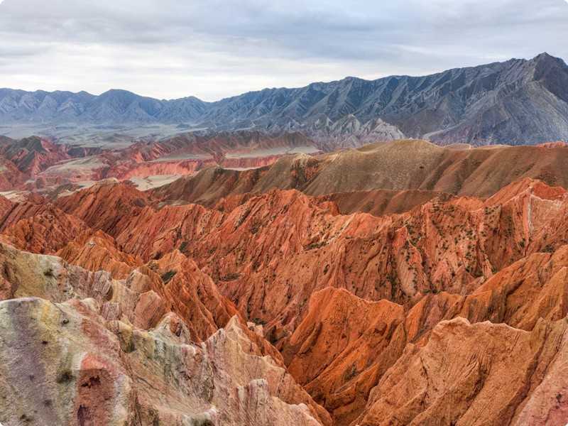 Paesaggio montuoso con vette arancioni in primo piano. Montagne bluastre si stagliano sullo sfondo sotto un cielo nuvoloso.