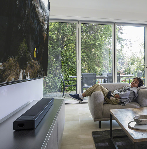 Soggiorno con TV, soundbar e divano. Una persona rilassata sul divano guarda fuori dalle grandi finestre. Tavolino da caff� in primo piano.