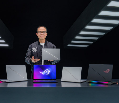 Uomo con giacca presenta un laptop. Sul tavolo, altri tre laptop di colore grigio e nero, con logo illuminato. Fondo nero.