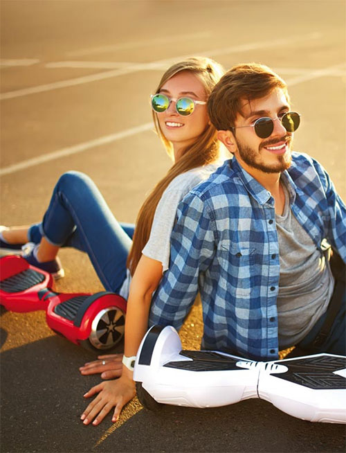 Uomo e donna seduti su hoverboard rossi e bianchi, indossano occhiali da sole. L'uomo indossa una camicia a quadri blu.