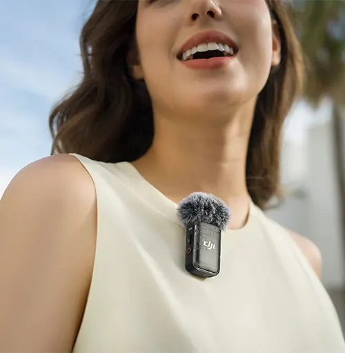 Donna sorridente con microfono clip-on grigio su una camicetta bianca. Cielo e vegetazione sullo sfondo.