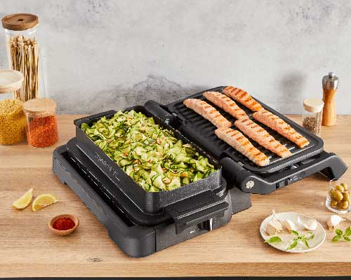 Griglia elettrica con zucchine e salmone. Sul tavolo, limoni, spezie, aglio e olive. Sullo sfondo, barattoli di pasta e spezie.