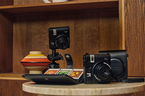 Due fotocamere nere su un tavolo con custodia, un vaso a strisce colorate e una scatola con immagini. Scaffale di legno sullo sfondo.