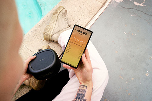 Persona tiene telefono e altoparlante neri. Telefono con schermo giallo. Persona con pantaloni bianchi e stivali accanto alla piscina.